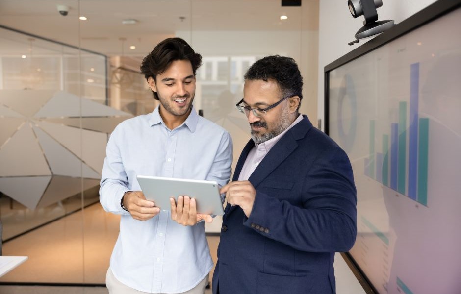 Dois homens de traje social discutem dados exibidos em um tablet em ambiente de escritório moderno, promovendo comunicação eficiente e análise de negócios.