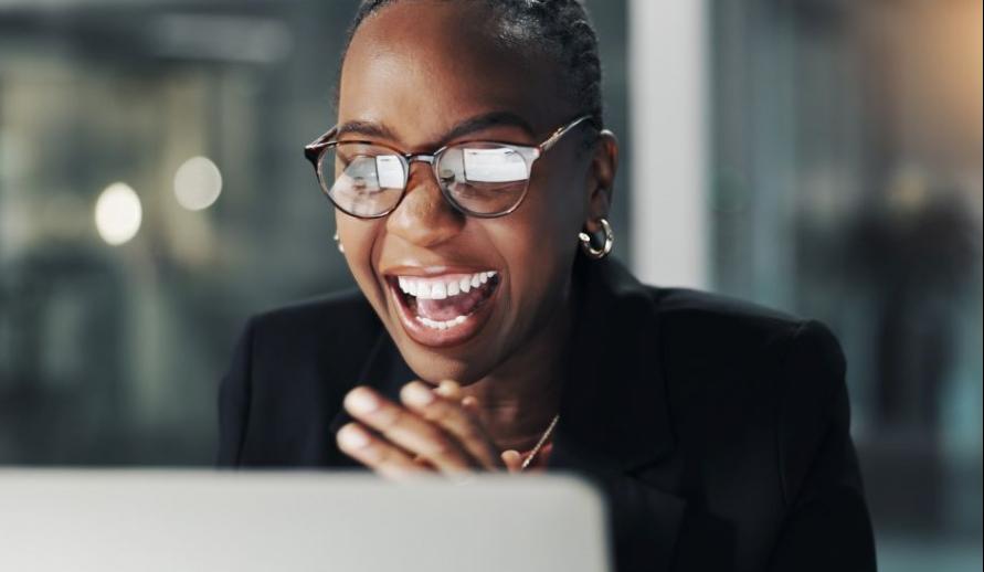 Mulher corporativa celebrando e sorrindo enquanto trabalha no computador, usando óculos e roupa elegante, representando sucesso, inovação e profissionalismo.