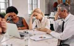 Equipe estressada em reunião, todos com as mãos na cabeça diante de relatórios e notebook, transmitindo frustração e sobrecarga.