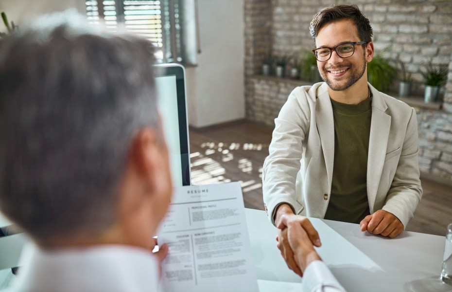 Homem de óculos sorri e aperta a mão do entrevistador, com currículo sobre a mesa, em uma entrevista de emprego.