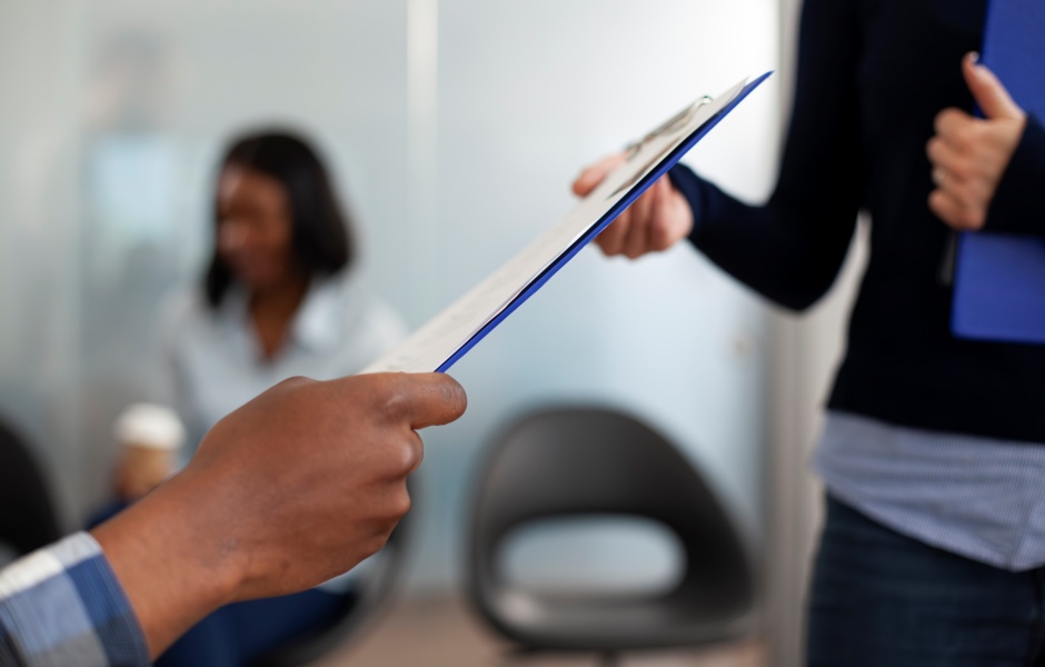 Mãos trocando uma prancheta com documentos em ambiente de escritório, sugerindo entrega ou revisão de formulários.
