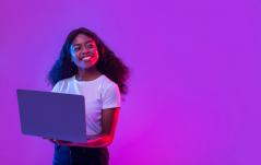 ma jovem negra sorridente segura um notebook aberto, iluminada por luzes vibrantes em tons de roxo e rosa neon.