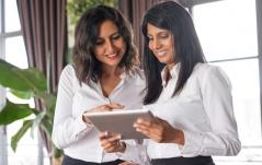 Duas profissionais sorridentes, uma morena e outra de traços indianos, analisam juntas o conteúdo de um tablet em um ambiente iluminado.