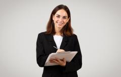 Mulher jovem com blazer preto sorri para a câmera enquanto faz anotações em uma prancheta branca, com fundo cinza neutro.