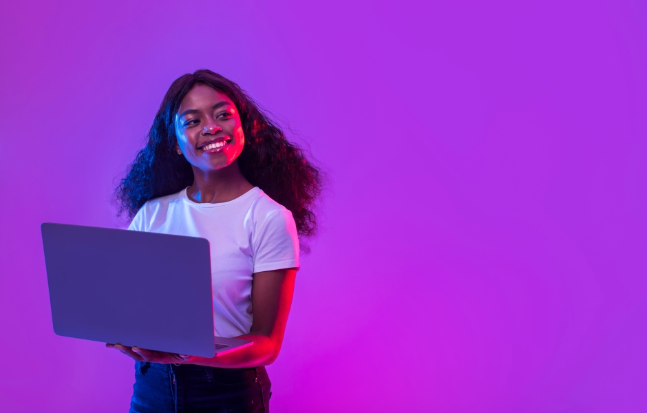 ma jovem negra sorridente segura um notebook aberto, iluminada por luzes vibrantes em tons de roxo e rosa neon.