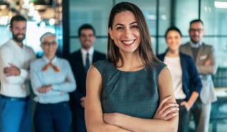  Mulher sorridente em traje formal cinza com braços cruzados, em destaque à frente de sua equipe.