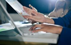Duas pessoas trabalham juntas, uma digitando no notebook enquanto a outra segura um relatório com gráficos.
