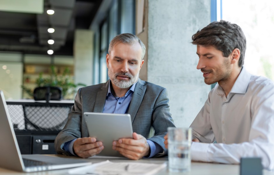 Dois homens de negócios analisam juntos um tablet em uma reunião, sugerindo mentoria ou discussão de resultados.