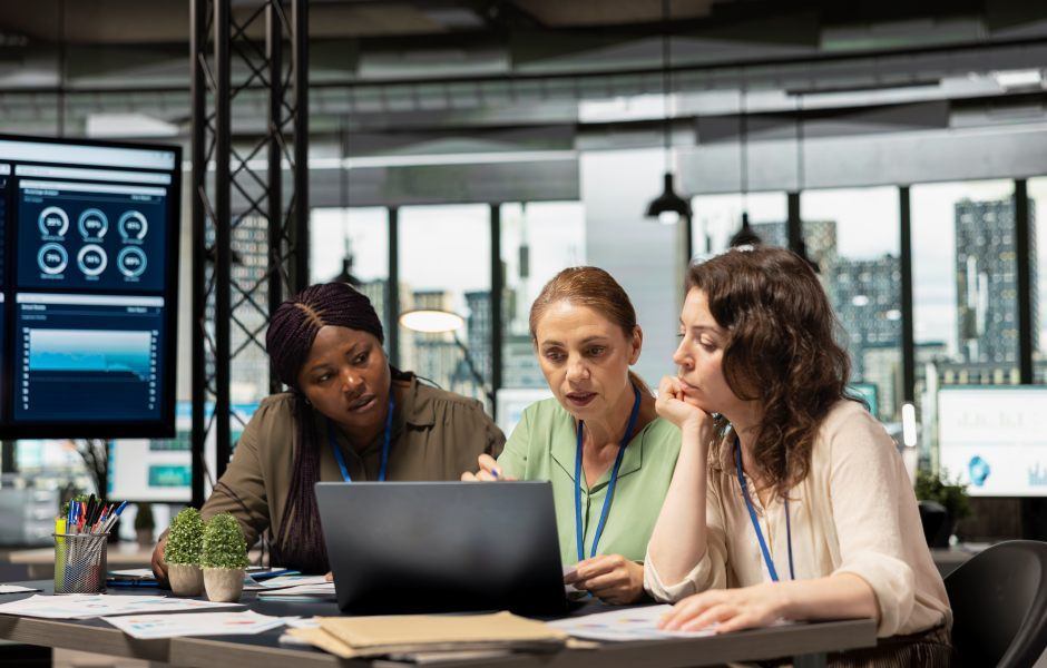 Três mulheres de diferentes etnias em uma reunião de trabalho, sentadas ao redor de uma mesa com um laptop, papéis e pequenos vasos de plantas.