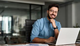 Homem jovem e barbudo, usando óculos e camisa azul, sorri sentado à frente de um notebook em um escritório moderno e organizado.