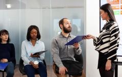 Grupo de candidatos sentados com pranchetas aguardando, enquanto uma profissional entrega documentos, sugerindo um processo seletivo ou triagem de candidatos.