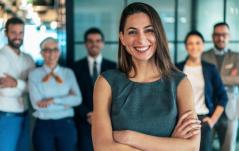  Mulher sorridente em traje formal cinza com braços cruzados, em destaque à frente de sua equipe.