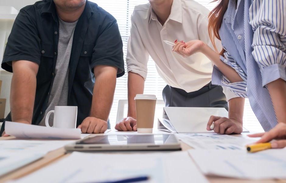 Três pessoas debruçadas sobre uma mesa com documentos, tablet e cafés, em uma reunião de trabalho informal.