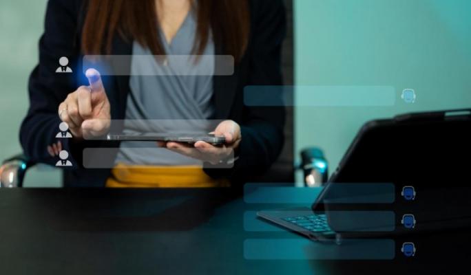 Profissional em escritório usando um tablet com interface de recrutamento/contatos sobreposta, com laptop aberto na mesa ao lado.