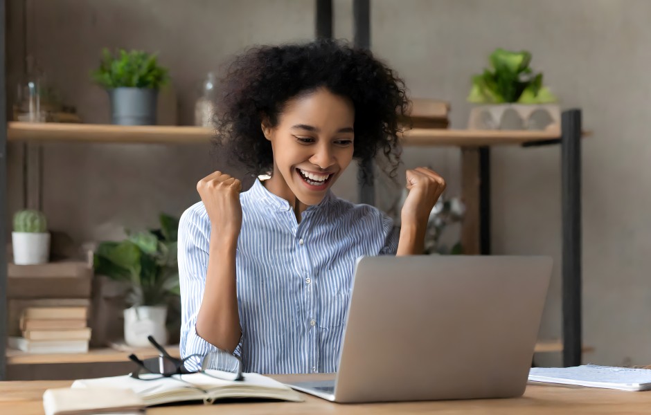 Mulher comemorando animada na frente do notebook, com punhos erguidos em sinal de vitória.