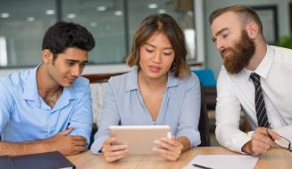 Três colegas analisam juntos um tablet em uma mesa, em ambiente de escritório colaborativo.