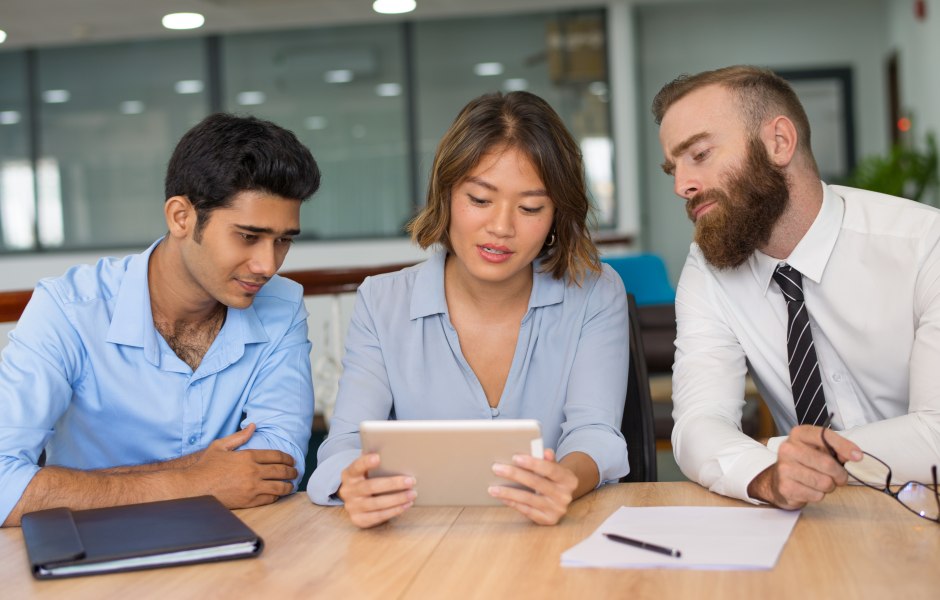 Três colegas analisam juntos um tablet em uma mesa, em ambiente de escritório colaborativo.