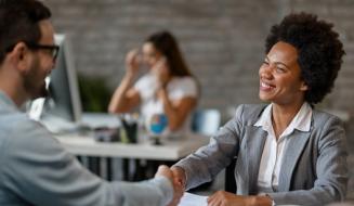 Mulher de terno cinza aperta a mão de um homem sorrindo, em um ambiente de escritório, sugerindo um acordo ou entrevista de emprego.