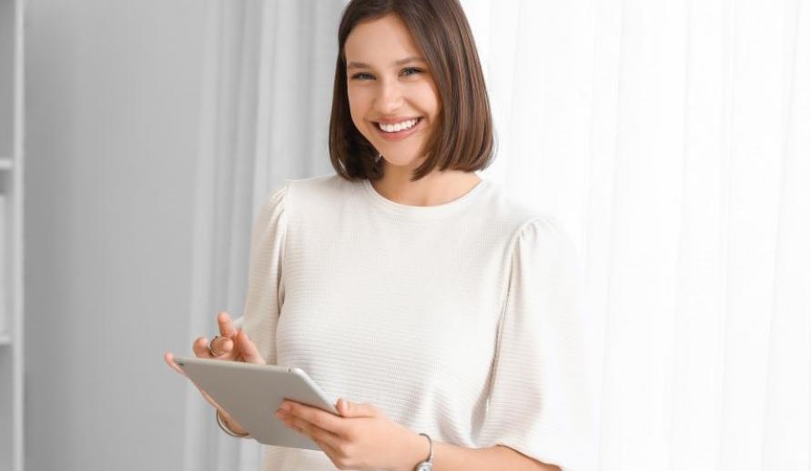 Mulher sorridente segurando um tablet em ambiente bem iluminado e moderno, transmitindo alegria e satisfação ao usar tecnologia.