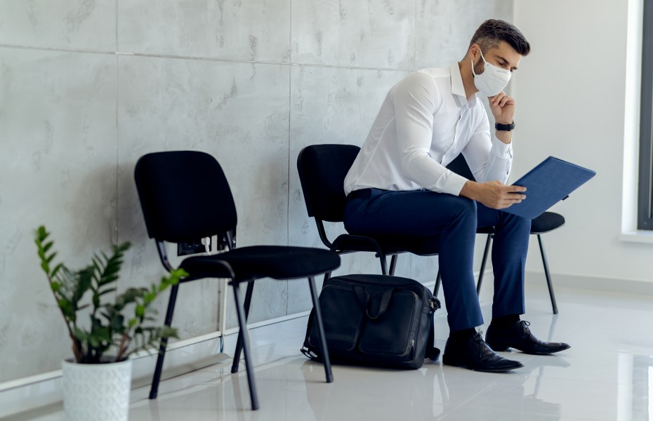 Homem de máscara e camisa social sentado em uma sala de espera, analisando atentamente documentos em uma pasta azul.