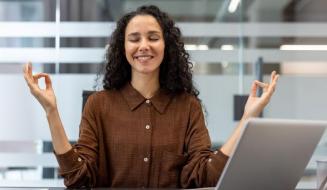 Mulher meditando com olhos fechados e sorrindo à mesa do escritório, transmitindo tranquilidade e equilíbrio no trabalho.