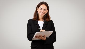 Mulher jovem com blazer preto sorri para a câmera enquanto faz anotações em uma prancheta branca, com fundo cinza neutro.