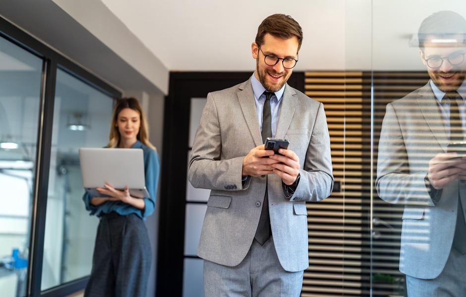 Homem de terno sorrindo ao mexer no celular, com colega ao fundo segurando um notebook.