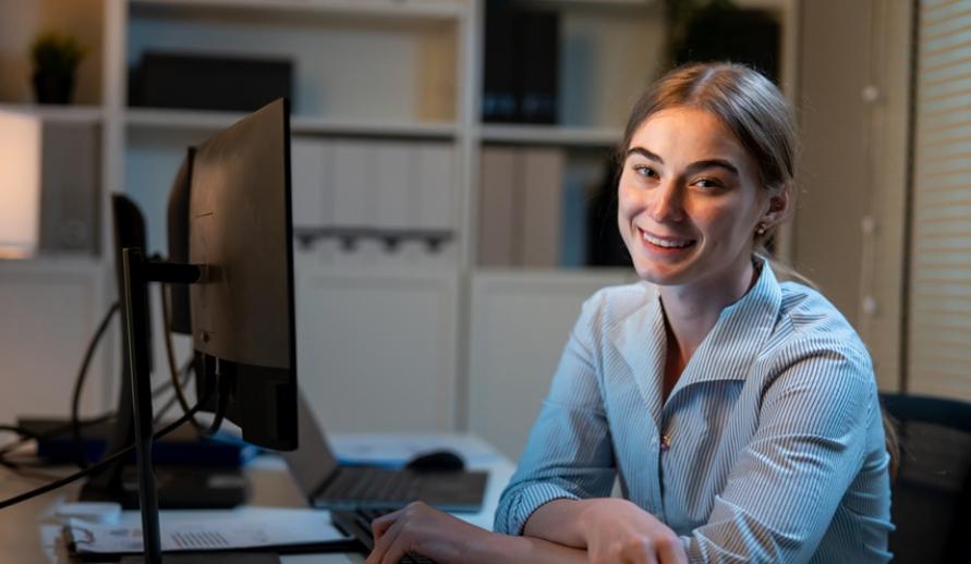 Mulher sorridente usando computador em escritório moderno, trabalhando de casa ou em ambiente profissional, representando produtividade e tecnologia.
