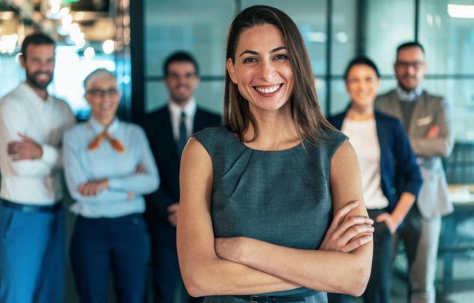  Mulher sorridente em traje formal cinza com braços cruzados, em destaque à frente de sua equipe.