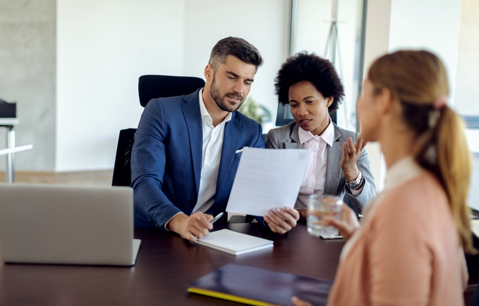 Close-up das mãos de uma pessoa segurando um currículo impresso com uma foto de perfil, enquanto outra pessoa ao fundo, levemente desfocada, observa a análise em um ambiente de escritório.