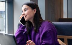 Mulher de moletom roxo fala ao celular sorrindo, sentada em um café ou escritório moderno.