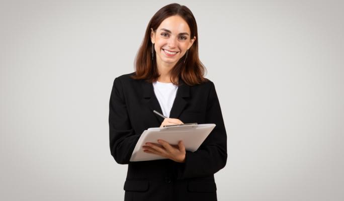 Mulher jovem com blazer preto sorri para a câmera enquanto faz anotações em uma prancheta branca, com fundo cinza neutro.