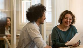 Dois colegas de trabalho, um homem com cabelo afro e uma mulher de óculos, conversam de forma animada e sorridente enquanto analisam um documento impresso em uma sala de escritório com divisórias de vidro.
