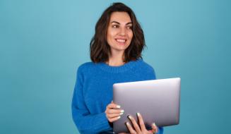 Uma jovem de blusa de tricô azul segura um notebook e olha para o lado com um sorriso alegre, sobre fundo azul.