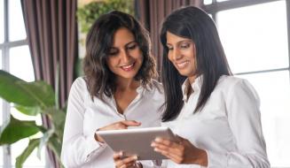 Duas profissionais sorridentes, uma morena e outra de traços indianos, analisam juntas o conteúdo de um tablet em um ambiente iluminado.