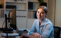 Mulher sorridente usando computador em escritório moderno, trabalhando de casa ou em ambiente profissional, representando produtividade e tecnologia.