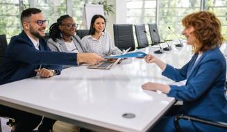 Grupo de pessoas em reunião corporativa ao redor de uma mesa branca, com um homem entregando uma pasta azul para uma mulher.