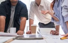 Três pessoas debruçadas sobre uma mesa com documentos, tablet e cafés, em uma reunião de trabalho informal.