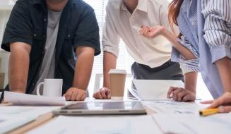 Reunião de equipe com três pessoas discutindo projetos em uma mesa de escritório, com café e documentos, ambiente corporativo profissional.