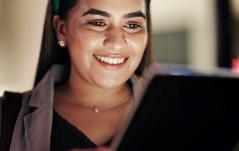Jovem mulher sorrindo enquanto usa um tablet, com a luz da tela iluminando seu rosto.