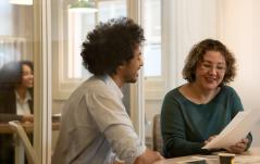 Dois colegas de trabalho, um homem com cabelo afro e uma mulher de óculos, conversam de forma animada e sorridente enquanto analisam um documento impresso em uma sala de escritório com divisórias de vidro.