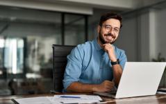 Homem jovem e barbudo, usando óculos e camisa azul, sorri sentado à frente de um notebook em um escritório moderno e organizado.