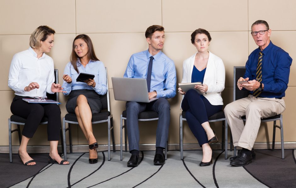 Cinco profissionais sentados em cadeiras de uma sala de espera, com laptops e pastas, demonstrando diferentes expressões de ansiedade ou conversa.