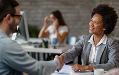 Mulher de terno cinza aperta a mão de um homem sorrindo, em um ambiente de escritório, sugerindo um acordo ou entrevista de emprego.