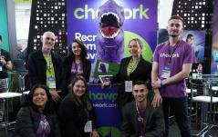 Grupo de sete pessoas da equipe Chawork posa sorridente em um estande de evento. Ao centro, destaca-se um mascote inflável de dinossauro roxo com a boca aberta, posicionado atrás de um balcão com o logotipo da empresa.