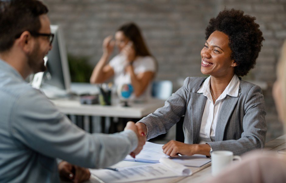 Mulher de terno cinza aperta a mão de um homem sorrindo, em um ambiente de escritório, sugerindo um acordo ou entrevista de emprego.