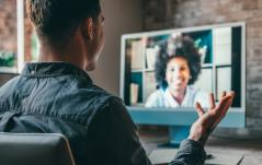 Homem em videochamada gesticulando para colega que sorri na tela do computador, representando uma reunião ou entrevista remota.