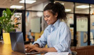 Jovem profissional sorridente de camisa azul trabalha em um notebook em um escritório moderno, com uma planta decorativa e documentos à sua frente.