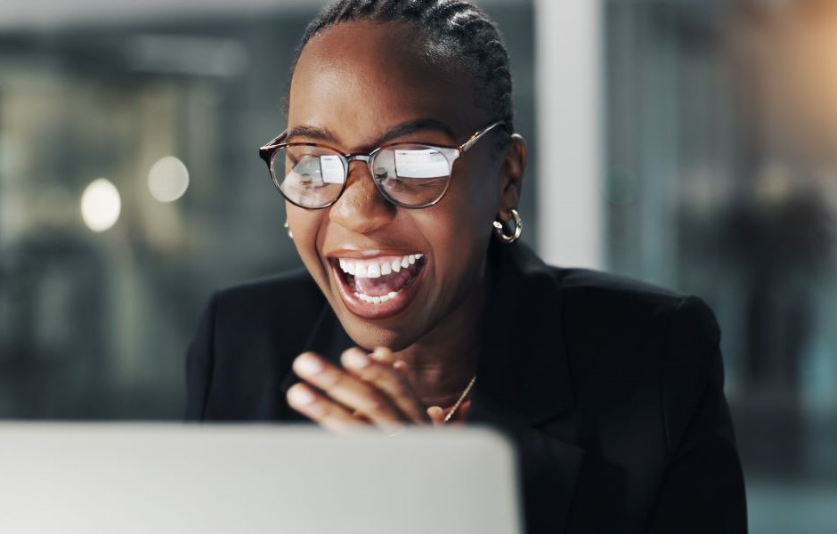 Mulher corporativa celebrando e sorrindo enquanto trabalha no computador, usando óculos e roupa elegante, representando sucesso, inovação e profissionalismo.