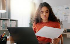 Jovem profissional analisa um documento com atenção em seu home office, com laptop aberto à frente e um mural de referências ao fundo.
